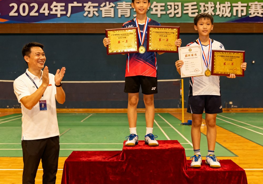 2024年广东省青少年羽毛球城市赛颁奖现场，CMP冠军国际学员站上领奖台，手持奖状和奖牌，教练在台下鼓掌祝贺。