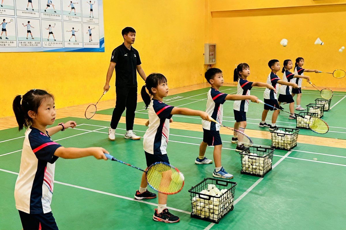 CMP冠军国际青少年基础班训练场景，一群6至8岁的小学员穿着统一训练服，在教练指导下认真练习羽毛球发球动作，每个孩子面前摆放着半筐训练用球，场馆墙面贴着基础动作分解图示。